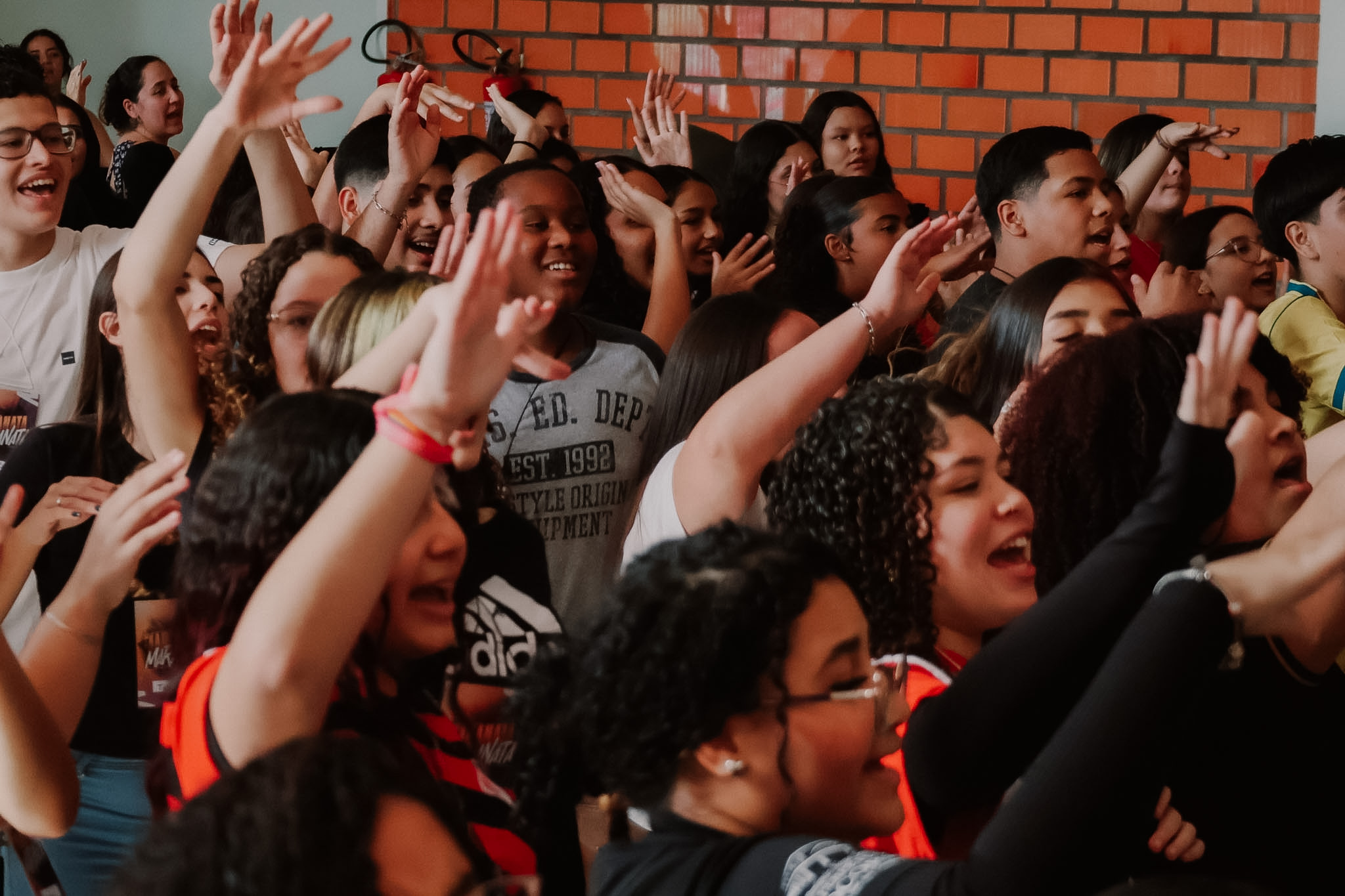 Dezenas de mãos levantadas no alto em um momento de louvor, diante de uma parede de tijolos